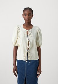Blusa di lino chiara con maniche a palloncino e chiusura con lacci sul davanti, abbinata a una gonna di jeans con spacco frontale.