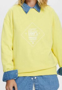 Žlutá mikina s grafickým motivem ve tvaru diamantu, na kterém je nápis „100% Organická bavlna“ v bílé barvě. Kontrast doplňují límec a manžety z denimu. Jemná textura.