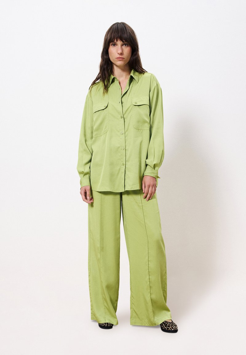 Lichtgroen, losvallend shirt met knoopsluiting en twee voorzakken, gecombineerd met bijpassende wijde broek. Zachte textuur en minimalistisch ontwerp.