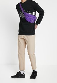 Lila crossbody-väska med dragkedjefickor, bärs över en svart sweatshirt med vita ränder, kombinerad med beige byxor och svarta sneakers.