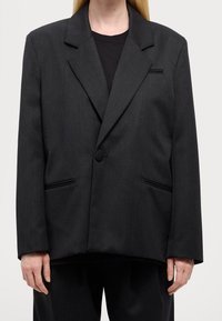 Czarna, dopasowana marynarka o zdecydowanym kroju. Charakterystyczne detale to szpiczaste klapy, dwie przednie kieszenie oraz pojedyncze zapięcie na guzik.