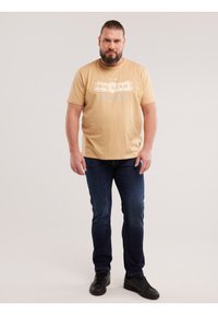 Bărbat cu barbă și părul prins la spate, purtând un tricou bej „Big Star”, blugi albaștri închiși și adidași negri, stând în picioare pe un fundal simplu.