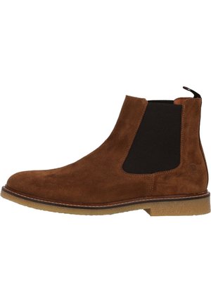 Bottes Chelsea en suede marron avec panneaux élastiques sur les côtés, lanière de traction et semelle en caoutchouc texturé. Des coutures soulignent la forme de la botte.