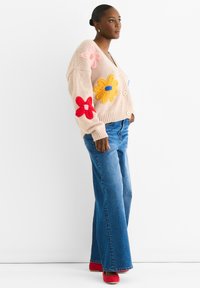 Light pink knitted cardigan with oversized, colorful flower appliqués. Paired with flared blue jeans and red shoes. Buttons on the front.