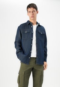 Denim shirt in dark blue with two chest pockets, button-up front, and a classic collar, worn over a white t-shirt and olive cargo pants.