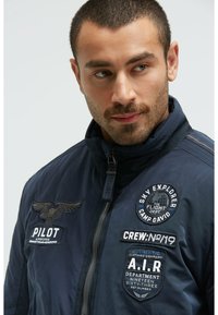 Dunkelblaues Bomberjacke mit Reißverschluss vorne, versehen mit mehreren gestickten Patches und einem Stehkragen. Texturiertes Material mit glatter Oberfläche.