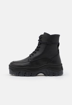 Botte en cuir noire montante avec semelle épaisse et robuste, et lacets à l'avant, montrée de profil sur un fond blanc uni.