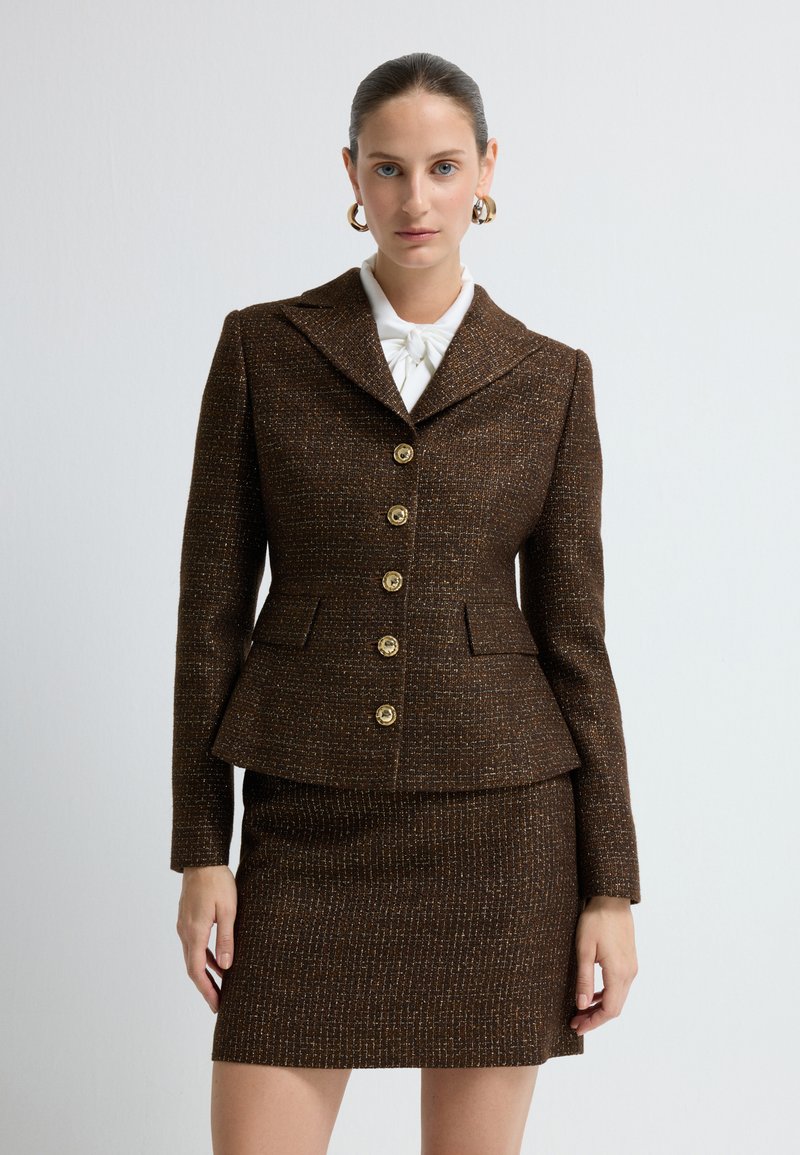 Brązowy garnitur z tkaniny tweedowej, składający się z dopasowanej marynarki z szpiczastym klapem, sześcioma złotymi guzikami oraz pasującą spódnicą. Teksturalny materiał z delikatnym wzorem.