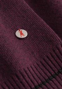 Bordeaux gebreide stof met een zilveren ronde tag met een rode hondensilhouet, bevestigd met een rode draad, met ribbeltextuur en detaillering.