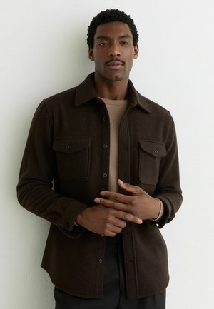 Man met kort krullend haar en snor, gekleed in een donkerbruine jas met knopen over een beige overhemd, staand tegen een witte achtergrond.