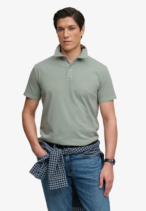Hombre joven con camiseta tipo polo verde claro, vaqueros azules, reloj, con camisa a cuadros azul atada alrededor de la cintura, de pie contra fondo blanco.