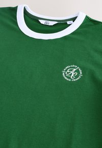 T-shirt verde in cotone con collo a girocostina bianco, caratterizzata da una stampa grafica circolare bianca con la scritta "ALPES-CÔTE D'AZUR" e dettagli di design.