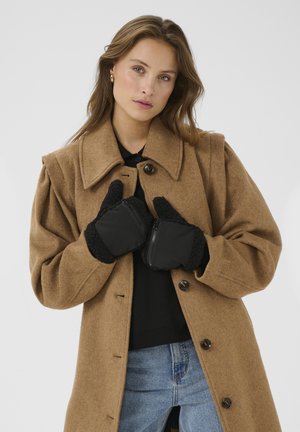 Brauner Wollmantel mit großem Kragen, schwarzen Knöpfen und aufgesetzten Taschen; schwarze Handschuhe mit Reißverschlussdetail und plüschigem Saum; das Model posiert mit Händen in den Handschuhen.