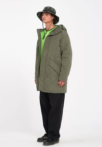 Olijfgroen gewatteerd jack met een capuchon, gecombineerd met een limoengroen shirt en zwarte broek. Zwarte schoenen maken de outfit compleet.