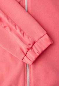 Gros plan sur la manche d'une veste rose corail avec un poignet élastique et une fermeture éclair argentée partiellement visible sur un fond en tissu assorti.