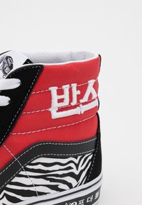 Tenis cu gheată înaltă, cu partea superioară din pânză roșie, accente din suede negru, model cu dungi zebra, text brodat și șireturi albe.