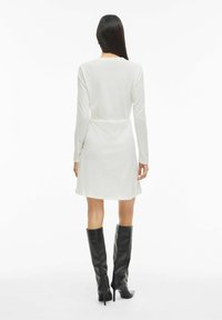 Robe blanche à manches longues côtelée avec une taille cintrée, une jupe évasée, et des bottes noires à talons montant jusqu'au genou ; texture lisse et design simple.