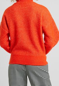 Osoba oblečená do jasně oranžového pleteného svetr a kalhot vzor houndstooth v černobílé barvě, zobrazená zezadu.