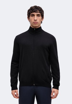 Homme aux cheveux courts et bouclés portant un pull noir à fermeture éclair intégrale avec col haut et pantalon foncé, debout devant un fond uni clair.