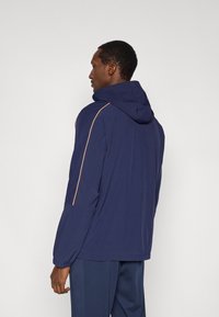 Ναυτικό μπλε μπουφάν με κουκούλα και πορτοκαλί λεπτομέρειες στους ώμους, με ελαφριά, λεία υφή και ρυθμιζόμενη κουκούλα. Η πίσω όψη δείχνει άνετη εφαρμογή.