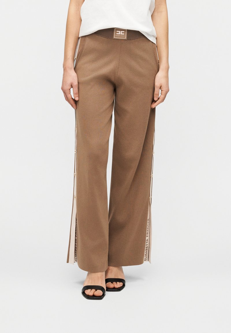 Καφέ παντελόνι με φαρδιά μπατζάκια και ψηλή μέση, με φερμουάρ στο πλάι και λεπτομέρεια λογότυπου στο τελείωμα. Συνδυάζεται με μαύρα σανδάλια ανοιχτής μύτης.