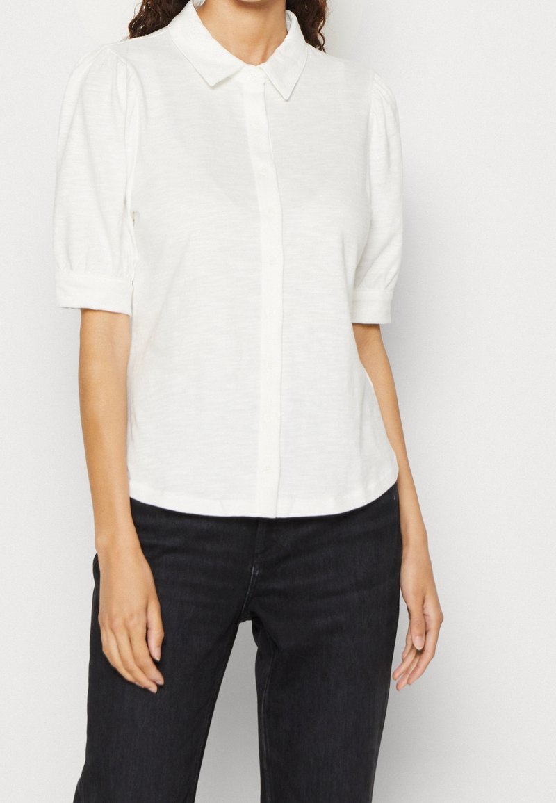 Blouse blanche à manches courtes avec un col et des fermetures à boutons, présentant une texture douce et une légère bouffante dans les manches.