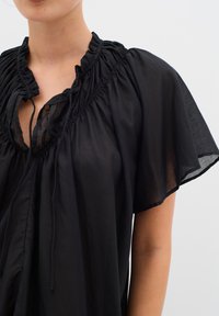 Top nero realizzato in tessuto leggero, con scollatura arricciata, maniche corte a farfalla e dettaglio con cordoncino al colletto.