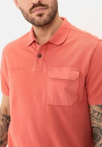 Koraalkleurig poloshirt van textured stof, met een placket met drie knopen en een borstzak links. Geborduurd "EXPLORERS" boven de zak.