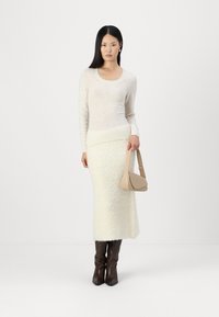 Suéter de punto color crema con escote redondeado, combinado con una falda midi blanca con textura y botas marrones hasta la rodilla. Lleva un bolso de hombro beige.