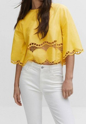 Kobieta w żółtym krótkim topie z okrągłymi wycięciami z koronki i falistymi brzegami, w połączeniu z białymi spodniami z wysokim stanem.