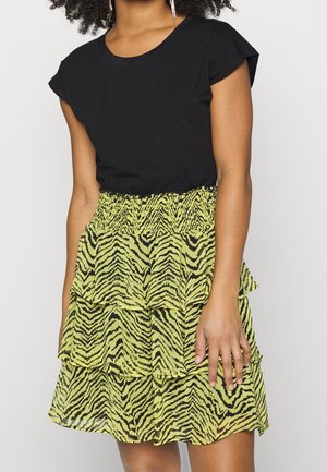 Woman wearing a black short-sleeved top and a yellow tiered skirt with a black zebra print, standing against a plain background.