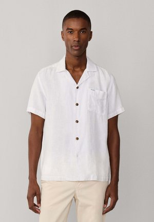 Hombre con camisa blanca de manga corta y botones, con un bolsillo en el pecho, combinada con pantalones beige claros, de pie frente a un fondo liso.