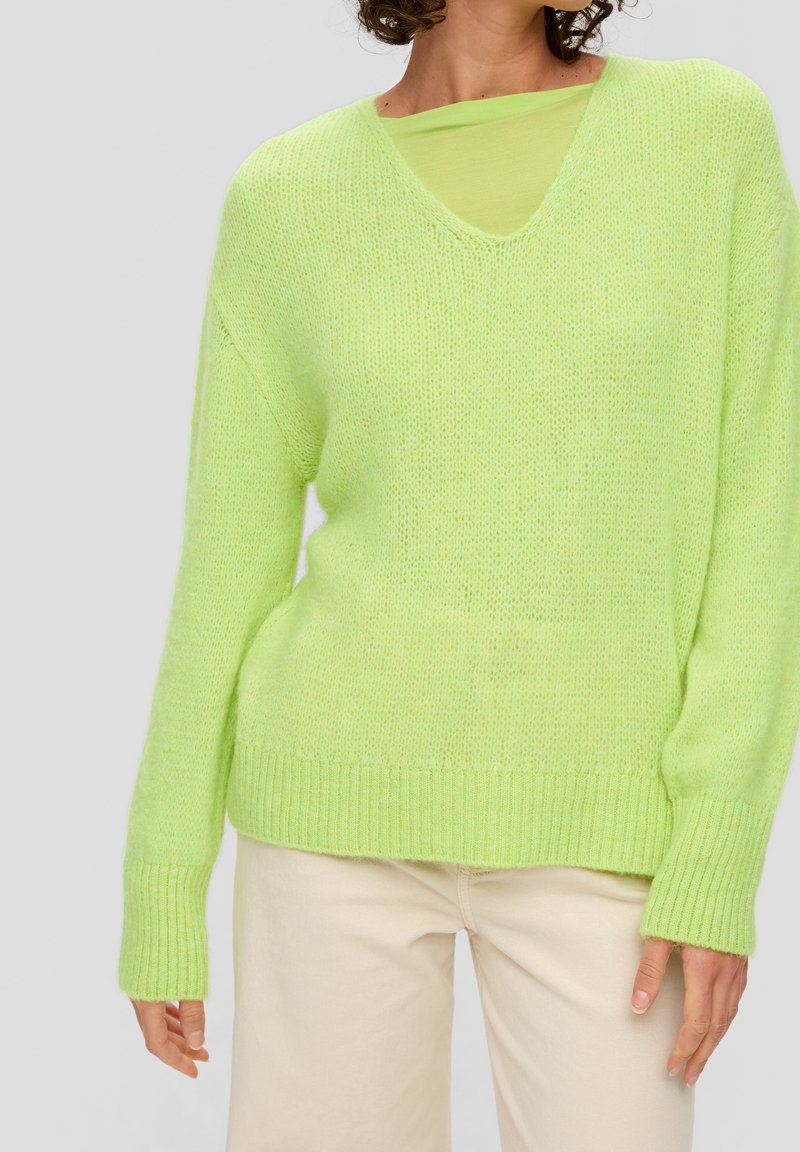 Femme portant un pull en maille vert citron vif à col en V par-dessus un haut transparent assorti, associé à un pantalon crème taille haute.