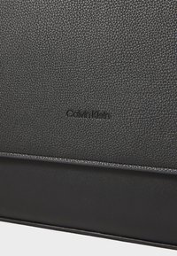 Borsa in pelle nera caratterizzata da una superficie testurizzata, logo "Calvin Klein" in rilievo e un pannello inferiore liscio. Design minimalista con linee pulite.