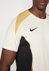 Camiseta deportiva de manga corta con un cuerpo blanco, acentos dorados en los hombros y un panel lateral negro. Logo de Nike en negro en el pecho.