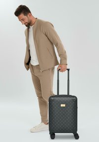 Valigia rigida nera con motivo a diamante testurizzato, targhetta con logo dorato, manico estensibile e quattro ruote, posizionata accanto a un uomo in abbigliamento casual.