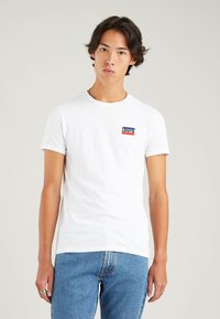 White cotton t-shirt with a crew neck, featuring a small red and blue Levi's logo on the left chest. Standard fit, smooth texture.