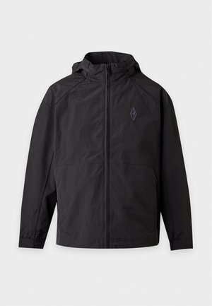 Un homme porte un bonnet noir, une veste, un pantalon et des bottes, avec une chemise blanche et un sac cabas en toile, devant un fond uni et clair.