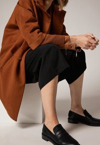 Abrigo de mezcla de lana color óxido con puños de canalé, combinado con pantalones negros cortos y mocasines de cuero negro con tacón plano.