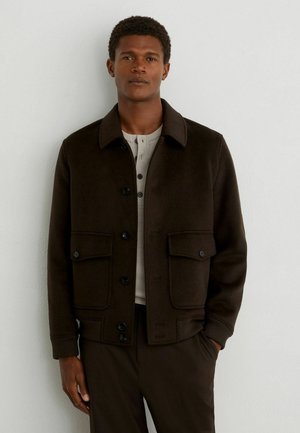 Uomo con capelli corti e ricci che indossa una giacca marrone scuro abbottonata sopra una maglia henley grigio chiaro, in piedi contro un muro bianco.