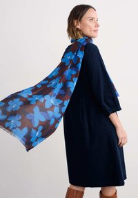 Blauer und brauner Schal mit großen Blumenmustern und fransigen Kanten, über einem dunklen Kleid drapiert, was einen geschichteten Look erzeugt.
