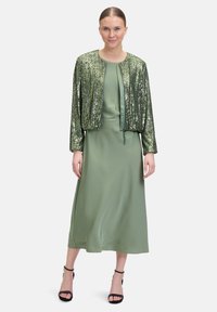 Grüne Paillettenjacke über einem glatten, knielangen grünen Kleid. Das Kleid hat einen klassischen Rundhalsausschnitt und eine fließende Textur. Schwarze Sandalen mit Knöchelriemen.