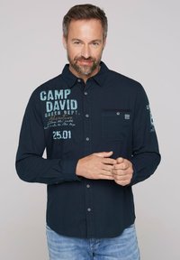 Camicia a maniche lunghe blu navy con chiusura a bottoni davanti, tasca sul lato sinistro del petto, e testo/design stampati sul davanti e sulla manica.