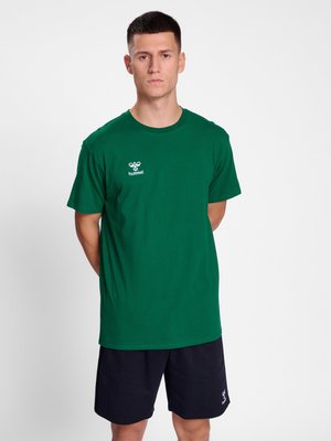 Ung mand stående med hænderne bag ryggen iført en grøn Hummel T-shirt og sorte shorts mod en ensfarvet hvid baggrund.