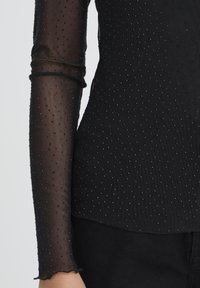 Top nero a maniche lunghe con tessuto trasparente a pois, caratterizzato da piccoli accenti metallici e orlo a scollatura. Il corpo è opaco con una texture morbida.