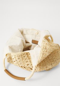 Sac seau en paille tissée, doublure en tissu beige, forme ronde, texture douce, avec des accents en cuir sur les poignées, fermeture à bouton sur le dessus.