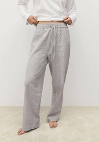 Szare i białe paskowane spodnie dresowe z elastycznym paskiem w talii, szerokimi nogawkami i miękką fakturą, leżące na bosych stopach na jasnym dywanie.