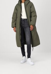 Abrigo acolchado largo de color verde oliva con capucha, cremallera frontal y un ajuste holgado, combinado con jeans oscuros y zapatillas blancas. Textura suave.