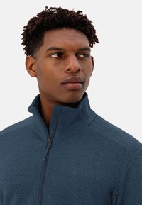 Giacca in pile con zip di colore blu navy caratterizzata da un tessuto texture, colletto alto e un logo discreto sul petto. Ingrandimento del collo e del viso.