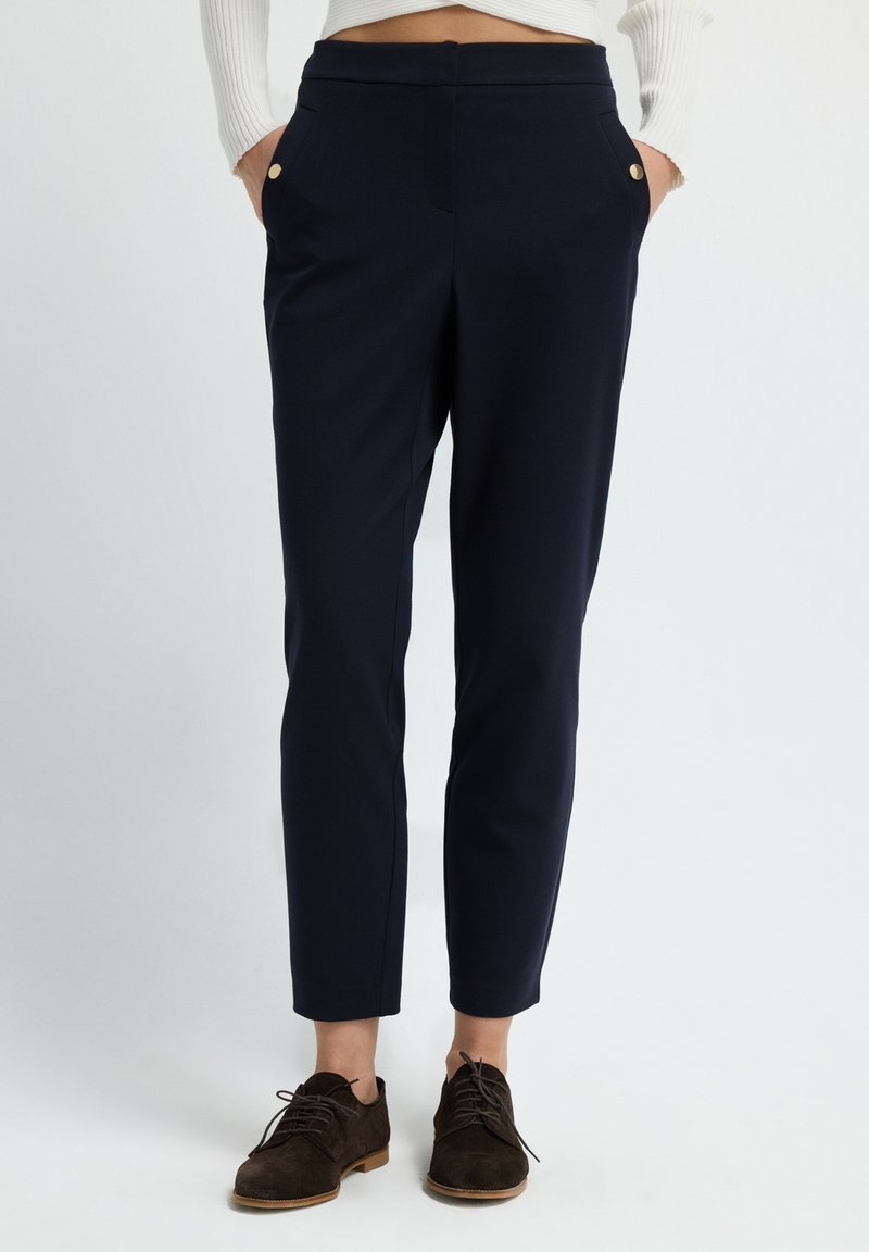 Donkerblauwe broek met een getailleerde pasvorm, voorzien van twee voorkbuiken, gouden knoopaccenten en een soepele textuur. Gecombineerd met donkerbruine schoenen.